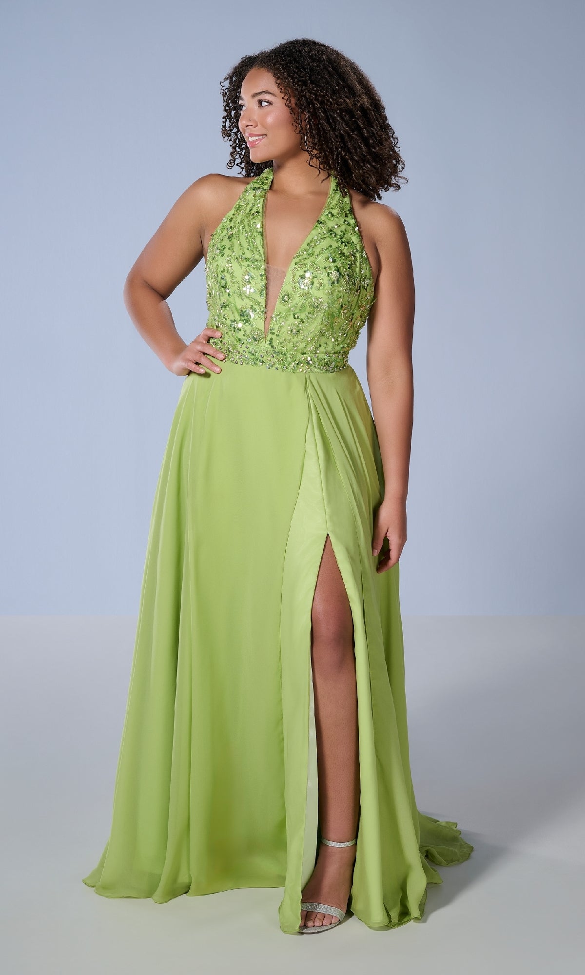 A woman stands against a plain background wearing the Tiffany 16167 long plus-size prom dress—a sleeveless, light green A-line gown with a sequin halter neckline, deep V-neck, and high front slit.